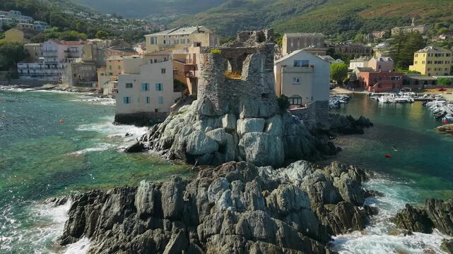 Erbalunga Cap Corse Corsica 4K drone video. Low altitude fly in over sea boulders to ruined Genoese tower rising up and pulling back to reveal authentic coastal village and mountains. 
