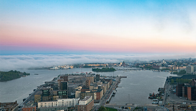 Aerial view of the Kalasatama district with fog rolling over the sea at sunset, featuring modern apartment buildings and harbor infrastructure in Helsinki, Uusimaa, Finland.