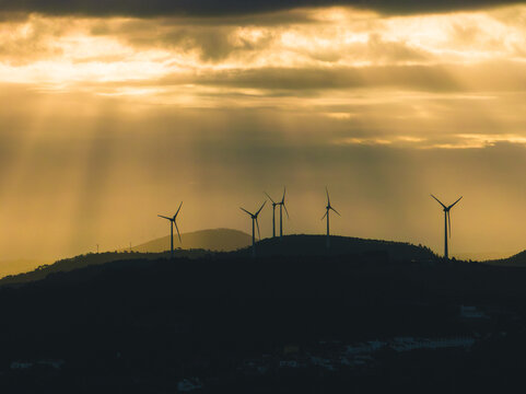 Aerial view of wind turbines on rolling hills at sunset with golden light rays breaking through clouds in Santo Isidoro, Lisboa, Portugal.