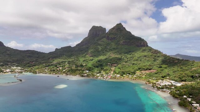 An aerial drone shot rises vertically over the deep blue lagoon, revealing a panoramic view of Mount Otemanu's rugged volcanic peaks and the lush green hillsides of Bora Bora