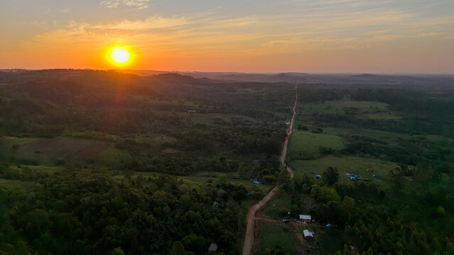 Aerial view of a rural landscape with a dirt road winding through green hills and dense forest under a vibrant sunset sky in Nueva Alborada, Itapua Department, Paraguay.