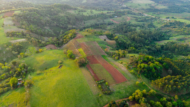 Aerial view of agricultural fields with red soil and green pastures surrounded by lush trees and rolling hills in Nueva Alborada, Itapua Department, Paraguay.