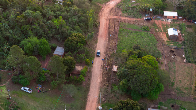 Aerial view of a rural dirt road winding through lush greenery and farmland with small houses and a car driving along the path in Nueva Alborada, Itapua Department, Paraguay.