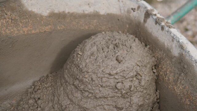 Close-up overhead view of thick wet concrete mortar forming rounded mound inside worn mixing container, with smeared gray walls, rough granular surface, soft blur behind, and natural even light