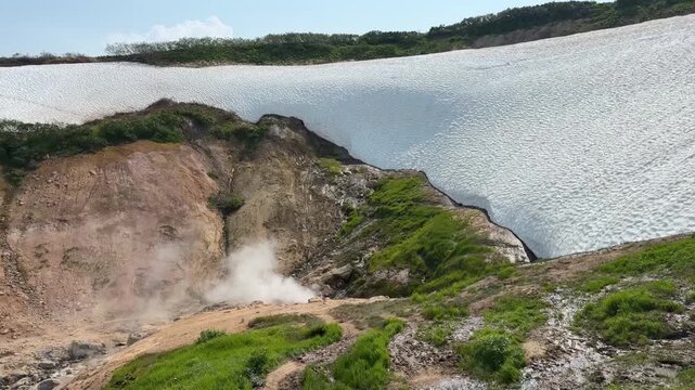 Steam rises from Dachnye Hot Springs beneath large summer snowfields and green slopes in Kamchatka. Geothermal activity, volcanic landscape, seasonal snowmelt, wilderness, nature tourism