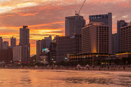A dramatic orange sunset glows behind dense high-rise buildings and construction cranes beside a crowded sandy beach, with warm light reflecting across the water.