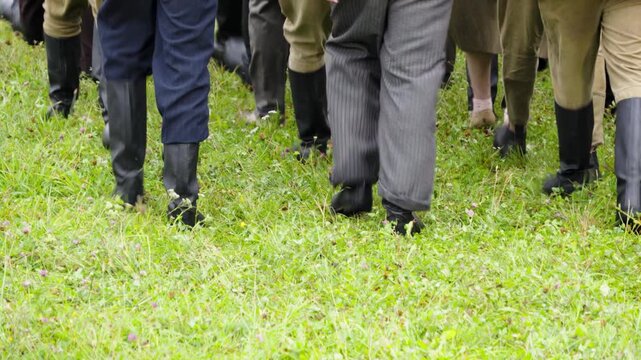 A large group of people wearing vintage military uniforms and period clothing march together in formation across a green grassy field during a historical reenactment event.
