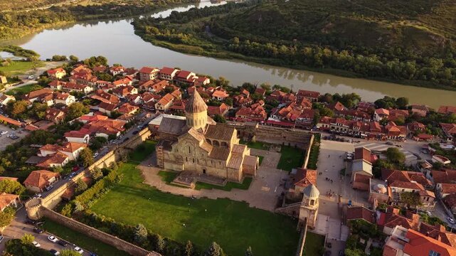 Drone slowly pulls away from Svetitskhoveli Cathedral, revealing a panoramic view of Mtskheta, the Caucasus mountains and Mtkvari River at sunset.