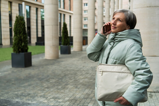 silver-haired businesswoman in her fifties is talking on a smartphone while standing next to a modern office building.