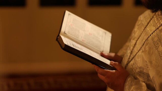 Hands holding open liturgical book over ornate brocade vestment during ceremony, with book, page, hand, vestment detail, showing brocade, embroidery, fabric, texture context