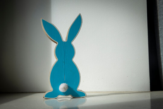 Blue wooden flat rabbit figurine with white pompom tail standing alone against white wall with dramatic side lighting and shadow.