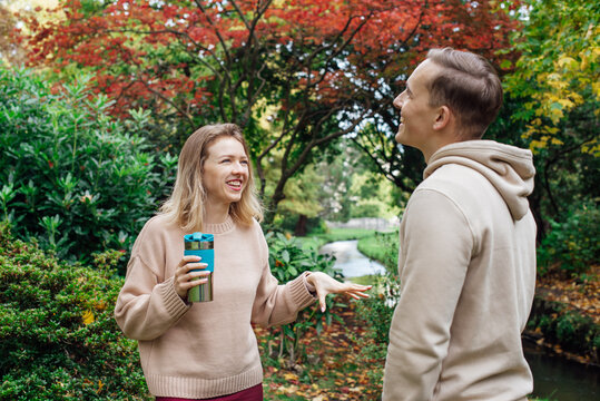 Two friends having emotional conversation outside in autumn park. They hold drinks in recyclable cups and smile at each other in a relaxed atmosphere during lunch or study break. Youth lifestyle