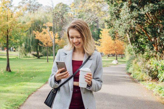 Smiling young woman using her phone while walking in autumn park for chatting, shopping, remote work, content creation for social media. Girl holding reusable cup with hot drink. and checking massages