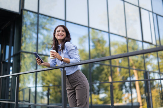 Businesswoman standing on balcony holding coffee and smartphone