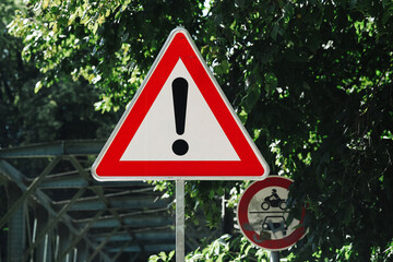 Obraz premium Red triangular warning sign with a black exclamation mark stands beside a road under green trees. General hazard symbol alerts drivers and road users to possible danger and caution background.
