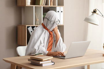 Ghost in a white sheet and orange tie working on a laptop at a wooden desk in a home office, the...