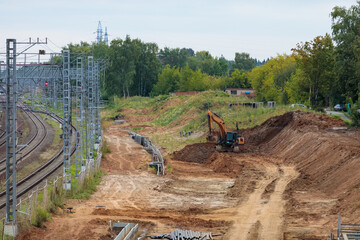 A hydraulic excavator operates on the embankment of a new railway construction site, leveling dirt near existing tracks and overhead power catenary lines. © ЮРИЙ ПОЗДНИКОВ