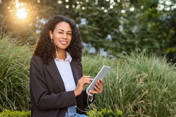 Fototapeta premium Remote work and digital nomad lifestyle, smiling woman using tablet with earphones in park during sunset for business communication and online learning