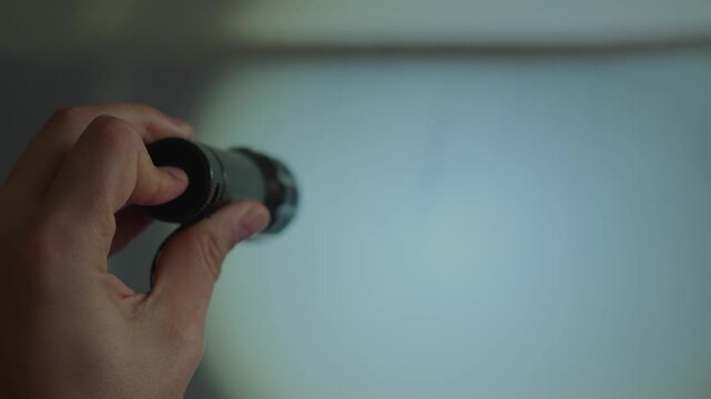 A hand holding and operating a compact black flashlight to illuminate a neutral gray surface. The flashlight alternates between continuous illumination and flashing strobe mode