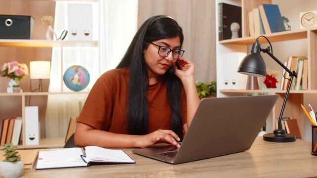 Indian woman on home table greets customer as support operator using headset and laptop for remote service. Arabian girl listens carefully and starts a friendly online client conversation from home.