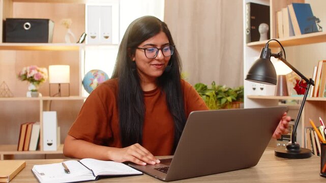 Indian woman on home table closes support request after solving client problem online and smiles at laptop. Arabian girl ends service call with calm expression and friendly wave after successful help.