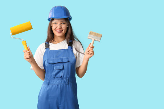 Young female painter with brush and roller on blue background