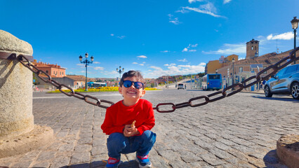 Cheerful young boy wearing sunglasses crouching beside a decorative chain in a sunny cobblestone plaza during a family trip in Spain © Sebastian Valencia P