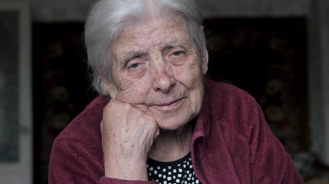 Thoughtful elderly woman with gray hair and deep wrinkles resting her chin on her hand, looking at the camera with a gentle and pensive expression, representing wisdom, longevity and nostalgia