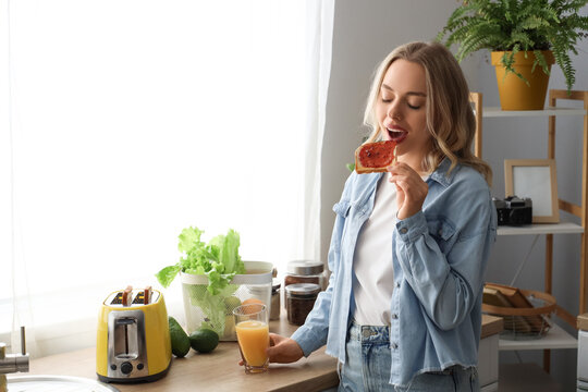 Young beautiful woman eating tasty toast with jam and drinking orange juice in kitchen