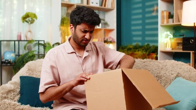Indian man on home sofa opens box to find wrong product with upset face from online order mistake indoors. Arabian guy on home sofa reacts to parcel problem with big angry hand gesture in living room.