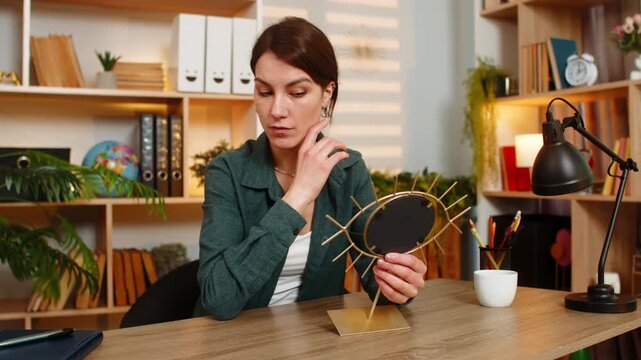 Young woman on home table enjoys mirror reflection smiles with joy during beauty self care break. Adult girl on home table feels happy adjusts earring checks appearance at home in living room indoors