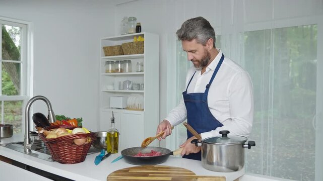 Man in apron making healthy food at kitchen. Cooking vegetables at home. Mature man cooking in home kitchen. Mature man preparing food for dinner. Old age man lifestyle in modern interior.