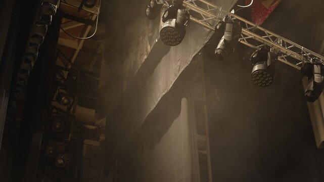 Video shows professional stage lighting rig with moving spotlights and led fixtures casting beams through haze in a dim theater space. Lights shift position creating dynamic mood