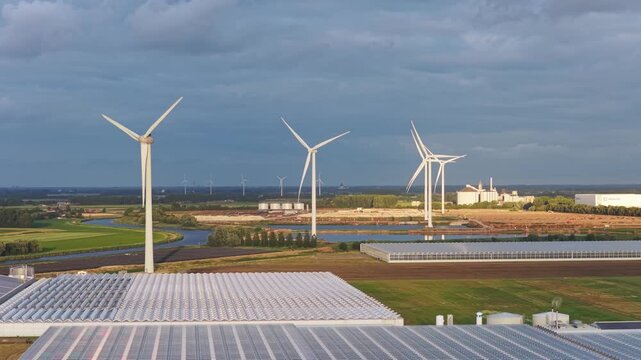 Aerial view of modern greenhouses for growing vegetables and fruits powered by wind turbines in the background. Sustainable green energy agriculture. 