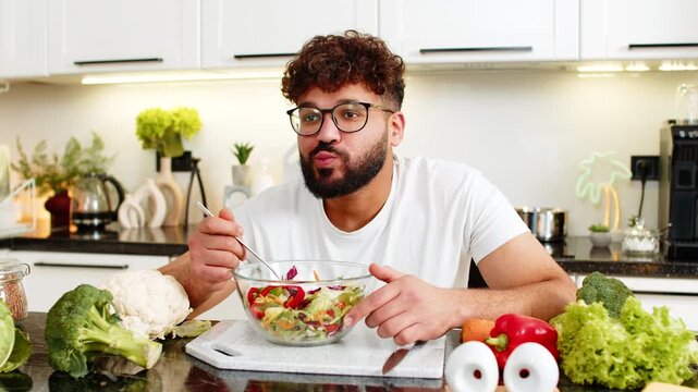 Arabian young man tastes homemade keto salad in home kitchen and eats with spoon smiling. Healthy nutrition concept showing low carb vegetables and fresh ingredients for diet fitness for wellbeing.