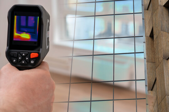 Hand holds a thermal camera. Its screen shows color-coded heat patterns. Building window with a grid forms the background. The person looks focused on the inspection task