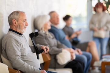 Fototapeta na wymiar Calm elderly man sitting in clinic waiting area with other senior patients in background