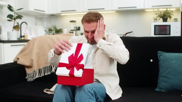 Young man at home opens present box lifts lid sees favorite item and gasps with warm awe and joy. Caucasian guy on sofa smiles holds gift close and shows joy surprise and grateful mood with soft vibe.