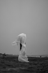 Person in white clothing stands on sandy beach during cloudy day. A person is standing on a sandy beach with a cloth blowing in the wind. The sky is gray. © Vadim