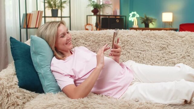 Pregnant middle-aged woman at home lies reading e-book on smartphone stroking belly smiling imagining motherhood. Caucasian girl on sofa enjoys fun story time bonding with unborn baby feeling pure joy