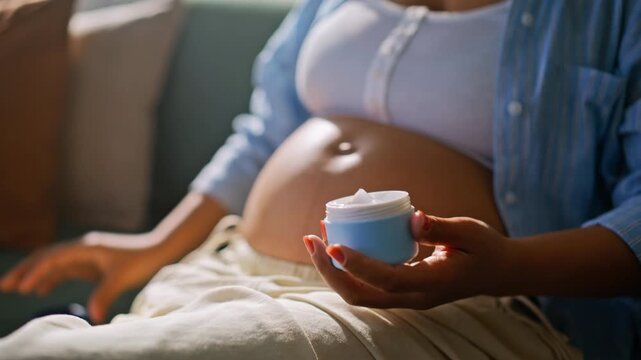 Pregnant woman scooping cream jar closeup. Mother moisturizing belly skin sofa.