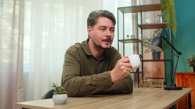 Middle-aged man at home sits with coffee cup enjoying quiet relaxing moment sipping warm drink peacefully. Young adult guy at table closes eyes feeling calm content comfort during gentle break today