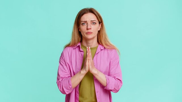 Middle-aged woman folds hands in front of chest with pleading expression, showing hope, gratitude or asking for help. Girl isolated on blue background uses prayer gesture to express request or emotion
