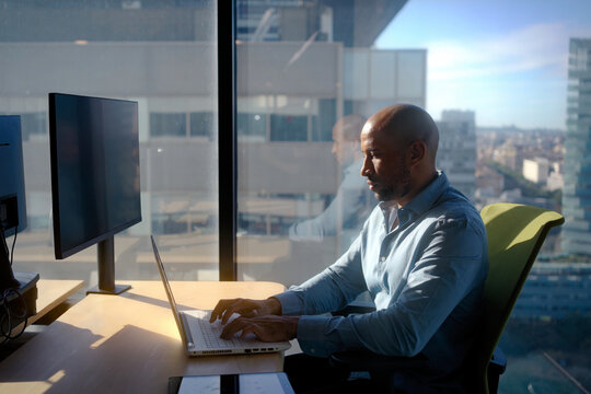 Sunlight streaming through expansive windows, revealing professional business African American man working intently on laptop within modern, sleek corporate office overlooking urban cityscape