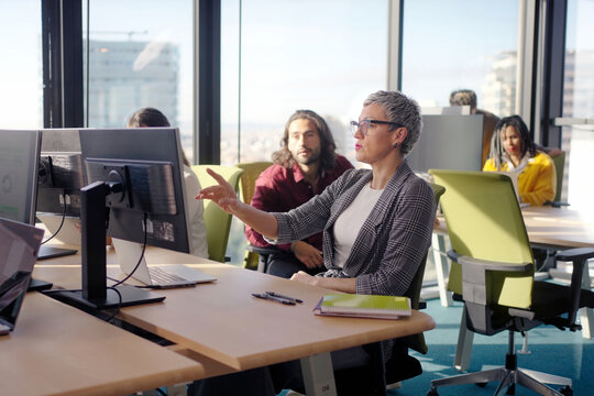 Confident mature businesswoman pointing at a monitor while explaining information to colleagues in a bright open office. Corporate scene conveying leadership, training and collaboration.