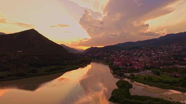 Cinematic drone flight above the Mtkvari River at sunset, showcasing the historic city of Mtskheta surrounded by mountains, rooftops and dramatic evening skies.