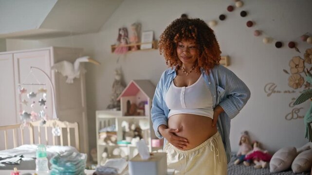 Smiling mother-to-be touching bare tummy standing nursery. Third trimester joy.