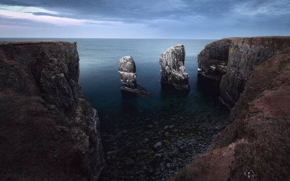 Dramatic sea stacks rise between rugged coastal cliffs at Elegug Stacks, Wales, under soft dawn light above calm dark water.