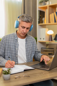 Middle-aged man on home table studies online course on laptop pauses video and writes notes in notebook. Young adult guy on home table repeats lesson and learns skills with focused calm mood indoors.