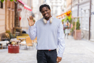 African American young man exploring city walking in city street and looking around with calm smile. Black guy tourist waves at camera outdoors during urban walk and cheerful city sightseeing break. © Andrii Iemelianenko